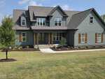 A charming two-story suburban house at 115 Water Willow Lane in Zebulon, featuring gray-green siding, wooden shutters, a covered front porch, and a newly landscaped lawn with fresh sod and small shrubs.