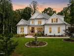 A large white two-story house with gabled roofs and many windows sits on 3511 Bruce Garner Road in Wake Forest, surrounded by green lawn, tall trees, and landscaping. A round stone fountain graces the front yard as the sky glows softly at sunset.