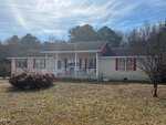 Charming single-story home at 44 Sutton Road, Louisburg, with light-colored brick, red shutters, and a cozy covered front porch. A bare tree and bush grace the front yard, framed by trees under a partly cloudy sky.