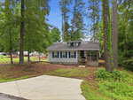 A small, single-story house with beige siding and blue shutters sits among tall trees. The house has a covered front porch and is surrounded by a natural yard with a concrete driveway in front.