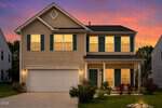 A two-story beige house with green shutters and a two-car garage at 837 Tannerwell Avenue, Wake Forest, is warmly lit from inside and sits against a colorful sunset sky. Well-kept shrubs and a small tree decorate the front yard.