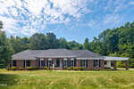 A single-story brick house with black shutters and white trim at 63 Opie Frazier Road in Henderson, set on a large green lawn with shrubs in front, surrounded by trees under a partly cloudy sky.