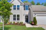 A two-story beige house with black shutters at 80 Silent Brook Trail, Franklinton, features a black front door with a wreath, an attached garage, and a landscaped front yard with shrubs and a tree on a sunny day.