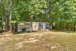 A small beige mobile home with white trim sits among tall green trees at 149 Rabbitt Lane in Henderson. The home features two windows, a cozy porch with stairs, and an American flag. The ground in front is bare, dotted with patches of grass.