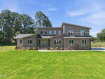 Modern two-story house with gray siding, large windows, and a double front door. The home sits on a well-manicured lawn with small shrubs and trees, under a clear blue sky.