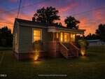 A small, green house at 2060 Julia Avenue in Henderson is warmly lit at dusk. Two bushes with ground lights flank the steps, and the sky glows orange and purple in the background.