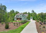 Nestled in Louisburg, 270 Whistlers Cove is a two-story green house with white trim and a covered front porch, set amid landscaped gardens and trees. A long driveway leads to a detached garage on the right under a clear blue sky.