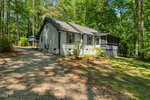 A small white house with a screened porch sits among tall trees on 956 Holden Road in Youngsville, surrounded by greenery. A gravel driveway leads up to the house, which is partially shaded by sunlight filtering through the trees.