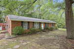 A single-story brick house with black shutters and a gray shingle roof sits surrounded by trees at 9531 Rueben Hart Road, Oxford. A small front porch with railings leads to the white front door, while stepping stones cross the mostly bare yard.