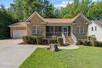Located at 2575 Mint Julep Drive in Creedmoor, this single-story brick house features a covered front porch with white railings, an attached two-car garage, a landscaped yard with a tree stump, and tall trees in the background.
