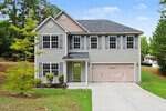 Charming two-story suburban house at 905 Woodland Road in Creedmoor, featuring gray siding, a double garage, multiple windows, and a covered entry. Enjoy the green lawn, small tree, shrubs, and tall trees in the background under a blue sky.