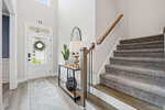 Bright entryway with a white front door, a glass side panel, a decorative wreath, a console table with plants and decor, a mirror above, and carpeted stairs with a wooden handrail leading upward.