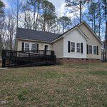 Charming single-story beige house at 121 Santee Drive, Louisburg, with black shutters and a dark roof. A wooden accessibility ramp leads to the front door. The home sits on a brick foundation, surrounded by tall trees and a grassy yard.