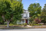 A white two-story Victorian-style house with a wraparound porch and a red front door sits at 305 N Main Street in Louisburg, surrounded by trees and greenery along a quiet street on a sunny day.
