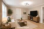 A bright, modern living room with light walls and wooden floors features a sofa, coffee table, TV on a wooden stand, potted plants, a side table with a lamp, and large windows with curtains. "Virtually Staged" text appears on the image.