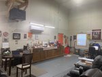 A garage or workshop with a wooden table and chairs, a long wooden cabinet, various tools, framed pictures, posters on the walls, and a recliner. There is a window, a red door, and equipment organized around the room.