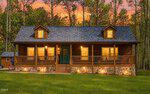 A cozy log cabin at 115 Running Bear Drive, Louisburg, with a large front porch glows warmly under a vibrant sunset sky. Stone accents line the foundation, and the house is surrounded by green grass and tall trees.