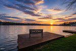 A decorative metal bench sits on a wooden dock overlooking a calm lake at sunset at 651 Sagamore Drive, Louisburg. The sky is partly cloudy with orange and purple hues reflected in the water. Grass borders the dock, and trees are visible across the lake.