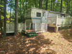 A white camper trailer with blue stripes sits elevated on blocks in a wooded area at 119 Geronimo Drive in Louisburg, surrounded by trees and pine-needle-covered ground, accessed by a ramp and stairs with white lattice railings.