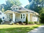 A light gray single-story house at 122 Military Street, Oxford, with a gabled roof, front porch with two chairs, white trim, and a neatly landscaped yard featuring small shrubs and trees on a sunny day.