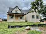 A newly constructed modern farmhouse at 1121 Dovefield Lane in Youngsville features gray siding, stone accents, wooden pillars, and a covered front porch on a grassy lot. Construction materials and erosion fencing are visible in the yard under a cloudy sky.