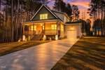 A warmly lit modern house at 158 Nakoma Drive, Louisburg, with a large front porch, wooden steps, and an attached garage, set against a wooded backdrop at sunset with a long driveway and neatly trimmed lawn.