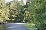 A paved road curves through a wooded area with dense green trees on both sides near 0 Heather Court, Franklinton. A mailbox and a boat on a trailer are visible to the left as sunlight filters through the leaves, casting shadows on the road.
