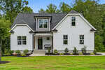 A modern white two-story house with a dark roof, multiple windows, a front porch, and neatly landscaped yard with green grass and shrubs, surrounded by trees.