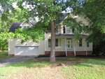 Two-story beige house with white trim at 1623 Rogers Pointe Lane, Creedmoor. Features include an attached two-car garage, a covered front porch, and a serene setting surrounded by trees and grass. A large tree in the front yard partially obscures the house, adding to its charm and privacy.