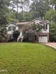 A two-story house with white siding, brick accents, and a garage located at 145 Huron Drive in Louisburg. A tree with pink flowers stands by the front steps. Lush green grass covers the yard, with tall trees and dense foliage in the background.