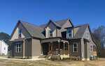 Two-story suburban house with brown siding, a covered front porch, and multiple gabled roofs sits at 15 Eagle Chase Drive in Youngsville. The yard is mostly bare with some small bushes near the porch. The sky is clear and blue.