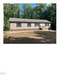 Single-story tan house with white trim and a front porch at 5586 Shelton Creek Road, Oxford, surrounded by trees and a spacious yard with a rock border near the house. Bright, clear weather with sunlight filtering through the green trees in the background.
