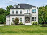 Two-story modern white house with black roof, double front doors, large windows, and a manicured lawn with bushes and trees, set against a backdrop of green trees under a partly cloudy sky in desirable Youngsville at 210 Scotland Drive.