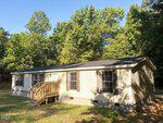 Charming single-story beige manufactured home with dark shutters and a small wooden porch, set on a grassy lot surrounded by tall green trees on a sunny day. Located at 116 S Creek Drive in Louisburg, this cozy residence offers peaceful living amidst beautiful natural surroundings.