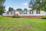Nestled on a grassy lawn at 174 Doctor Finch Road, Henderson, this long, single-story manufactured home features beige siding, black shutters, and a brick foundation, with a few small shrubs and trees providing a serene backdrop.