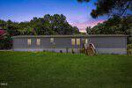 A single-wide gray mobile home with lit windows and a small front porch sits on a large grassy lawn, bordered by trees, at dusk under a purple and blue sky at 485 Lemay Lane in Henderson.