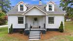 White house with two dormer windows, a covered front porch, black railings, and steps leading to a white front door with a wreath. Two small shrubs and flower pots are near the entrance. Green lawn and trees surround the house, offering inviting curb appeal at 5083 NC 56 Hwy in Franklinton.