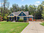 Charming blue house with white trim at 1212 Rogers Farm Road in Wake Forest, featuring a welcoming front porch, attached garage, and well-kept lawn. Surrounded by trees, there's a clear driveway leading up to the house under a bright, partly cloudy sky.