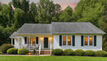 A white single-story house with black shutters, a front porch, and lit windows sits amid green bushes and trees at sunset on 104 Meadow Lane in Zebulon.