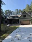 A two-story house with a covered front porch and attached garage sits among tall pine trees. The driveway is concrete with scattered pine needles, and the sky is clear and blue.