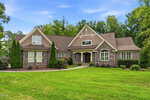 A large, two-story house with stone and wood siding, tall peaked roofs, arched windows, green lawn, and shrubs, set against a backdrop of trees under a blue sky at 1106 Dovefield Lane in Youngsville.