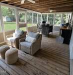A screened-in porch at 104 Pineview Drive, Louisburg features wooden floors, two wicker armchairs, pouf ottomans, a dining table with chairs, and ceiling fans. Large windows offer a view of a green backyard and shed.
