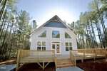 A modern white house with large windows, a wooden porch, and steps sits gracefully at 253 Sandy Point Drive, Manson, surrounded by tall trees in a forested area under a clear blue sky.