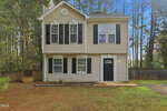 Two-story beige house with black shutters, white trim, and a black front door located at 60 Holding-Young Road in Youngsville. Surrounded by tall pine trees and a wooden fence, this home features a simple front yard and a small garden bed near the entrance.