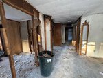 Interior of a run-down house at 414 Spring Court, Henderson, with exposed wall studs, broken drywall, debris scattered across the floor, and a large trash bin filled with construction waste in the center. Light comes in from the left.