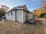 Small beige house with a boarded-up door, wooden ramp, and overgrown grass yard at 410 Spring Court. Autumn trees and a driveway with a car are visible in the background under a clear sky in Henderson.