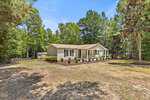 Single-story beige house with a front porch, white railings, and flower beds, nestled among trees on a spacious yard at 175 Windsong Lane in Youngsville, all beneath a clear blue sky.