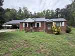 A single-story brick house with a metal roof, front porch steps, and a covered carport on the left sits at 523 US Highway 158 in Rougemont. The yard is grassy with some patches, and trees line the background under an overcast sky.