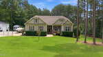 Single-story house with beige siding and green trim, surrounded by a large, neatly mowed lawn, tall trees, and shrubs. A boat is parked on the driveway beside the house, with a forested area in the background.