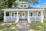 Charming light green house with white trim at 503 Kenmore Avenue, Louisburg. Features a wide front porch and classic brick columns. A welcoming path leads to the front door, surrounded by trees and neighboring houses. Enjoy the blue sky with a few clouds in the background.